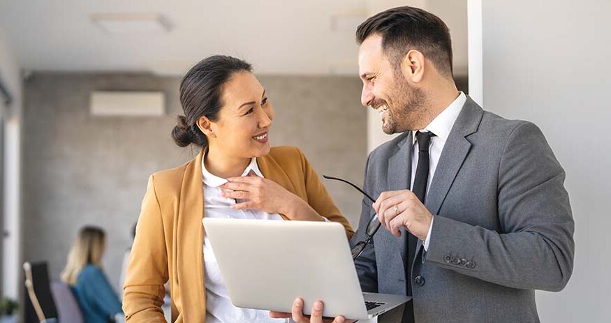 A woman and man in business wear standing next to each other with a smile on their faces and the man on the right holding a laptop in his right hand