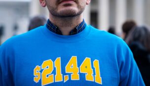An image of a man's lower part of his face and chest as he wears a blue sweatshirt with "$21,441" printed on it in yellow