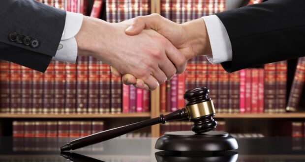 Two people shaking hands with law books in the background and a table with a gavel on it on a table below the hands